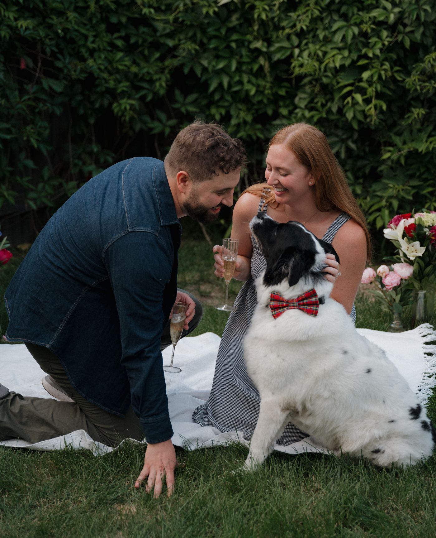Lauren and Jonathan laughing together outdoors with their dog, who is wearing a plaid bow tie